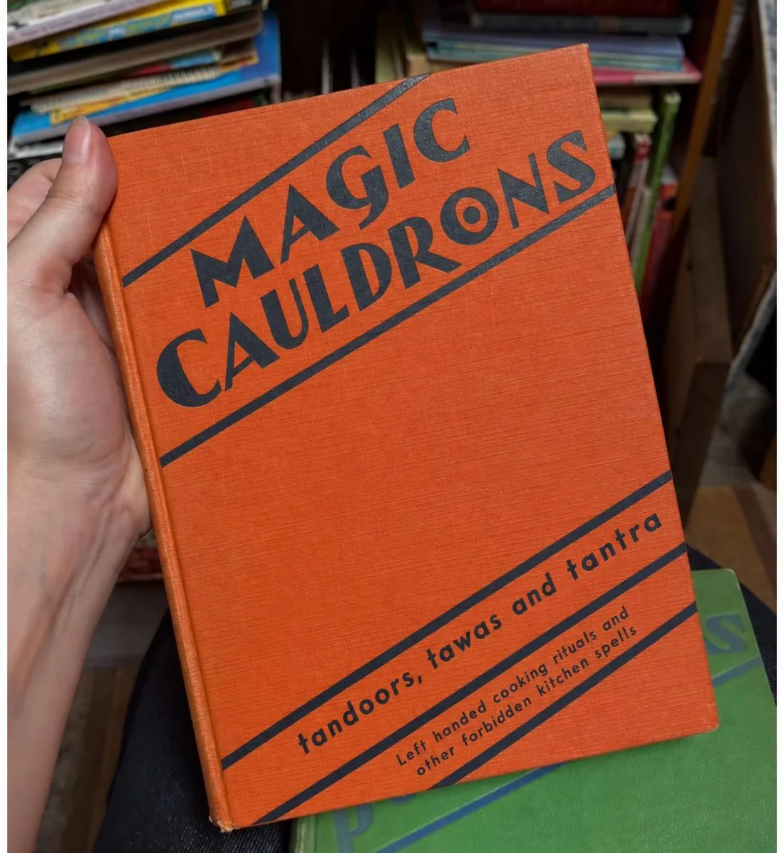 Orange cloth-bound book held in a hand: Magic Cauldrons. Tandoors, tawas and tantra. Left handed cooking rituals and other forbidden kitchen spells.
