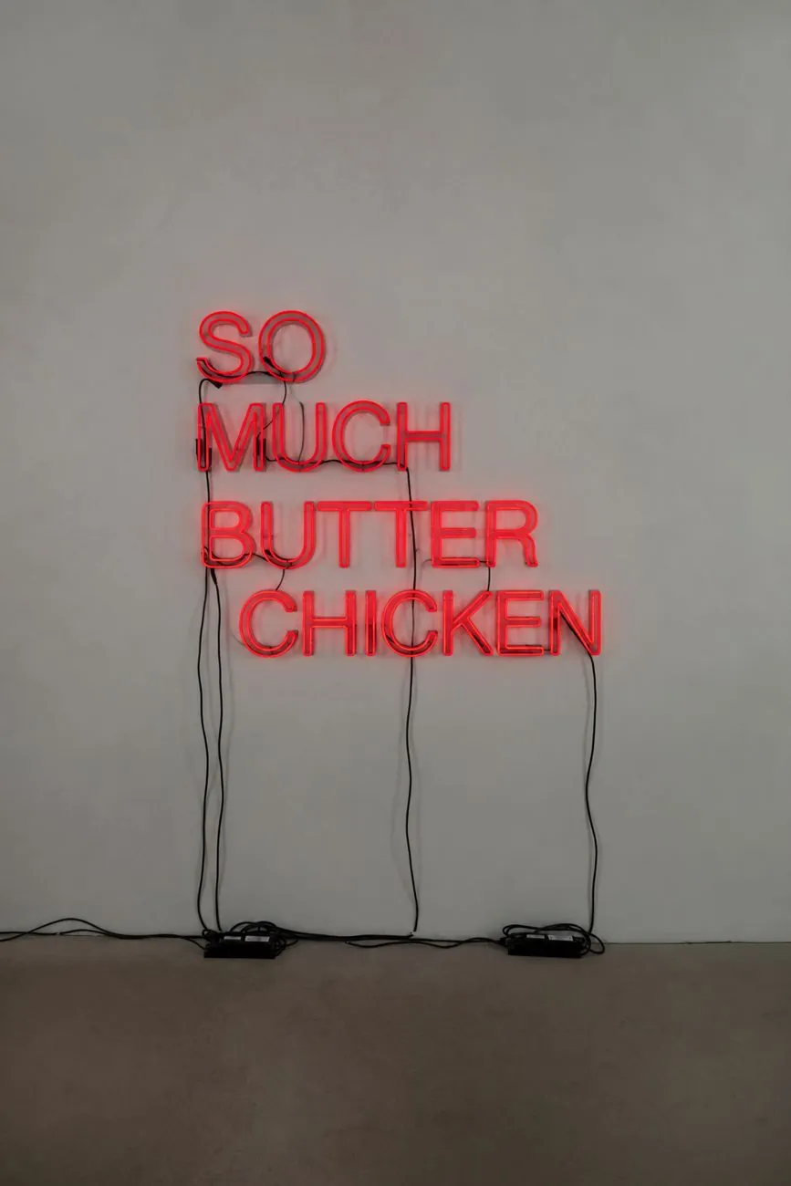 Red neon sign on a grey wall reading So Much Butter Chicken; the word Chicken flickers on and off
